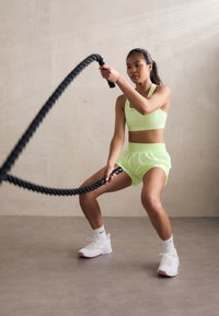 Neon zelena sportska grudnjak i kratke hlače, crna borbena uže, bijele atletske cipele s ružičastim detaljima, u fitness studiju s neutralnim zidovima.