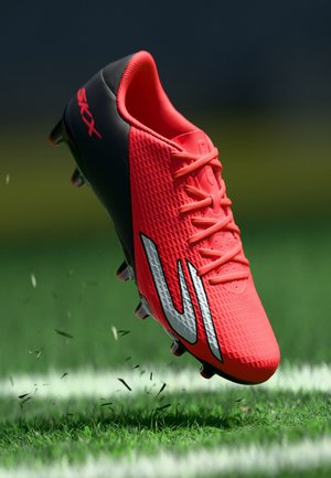 Klar rød fodboldstøvle med sort hæl og sølvlogo, der svæver over græs på en markeret sportsbane.