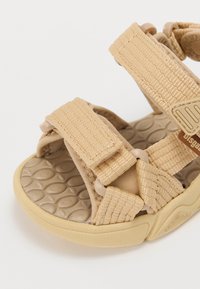 Beige Sandalen mit verstellbaren Stoffriemen, strukturiertem Gummisohle und geformtem Fußbett. Sie verfügen über ein rundes, leichtes Design.