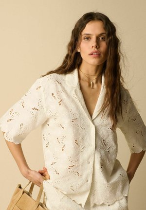 Vrouw met lang golvend haar draagt een witte eyelet blouse en ketting, houdt een beige handtas vast tegen een neutrale achtergrond.