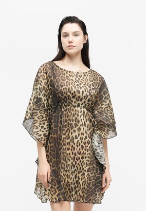 Nő, aki laza, áttetsző, leopárdmintás ruhát visel, széles ujjakkal, egyszínű fehér háttér előtt áll.