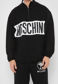 Osoba oblečená v černém svetru Moschino s bílým logem na pruhu a odpovídajících černých kalhotách s bílým logem Milano na stehně.