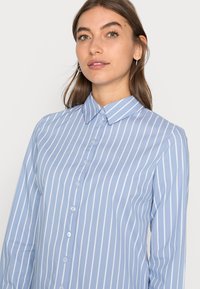 Camicia azzurro chiaro con righe verticali bianche, colletto strutturato e maniche lunghe. Presenta un tessuto liscio e sette bottoni.
