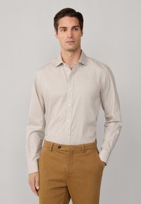 Camisa abotonada de color beige claro con un patrón a cuadros, combinada con chinos marrones. Tejido de algodón, cuello estructurado, mangas largas.