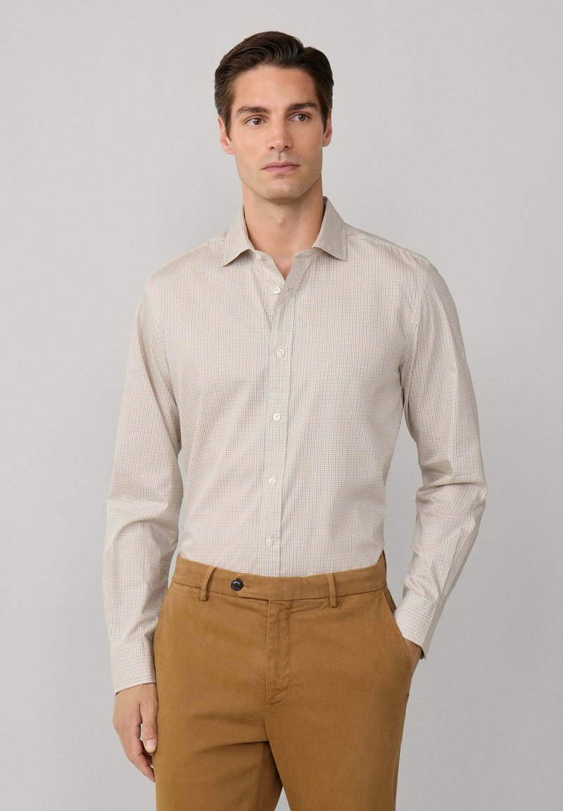 Camisa abotonada de color beige claro con un patrón a cuadros, combinada con chinos marrones. Tejido de algodón, cuello estructurado, mangas largas.