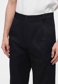 Homme portant un pantalon noir sur mesure avec une fermeture à boutons et une chemise blanche rentrée, debout les mains détendues le long du corps.