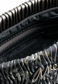 Interior de bolso de cuero negro texturizado con diseño plisado, que presenta un acento de logo plateado y un patrón floral gris en contraste. Compartimento con cremallera visible.