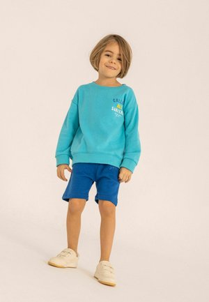 Ljus turkos sweatshirt med en logotyp, kombinerad med blå shorts. Barnet har på sig vita sneakers, vilket visar en avslappnad, casual outfit.