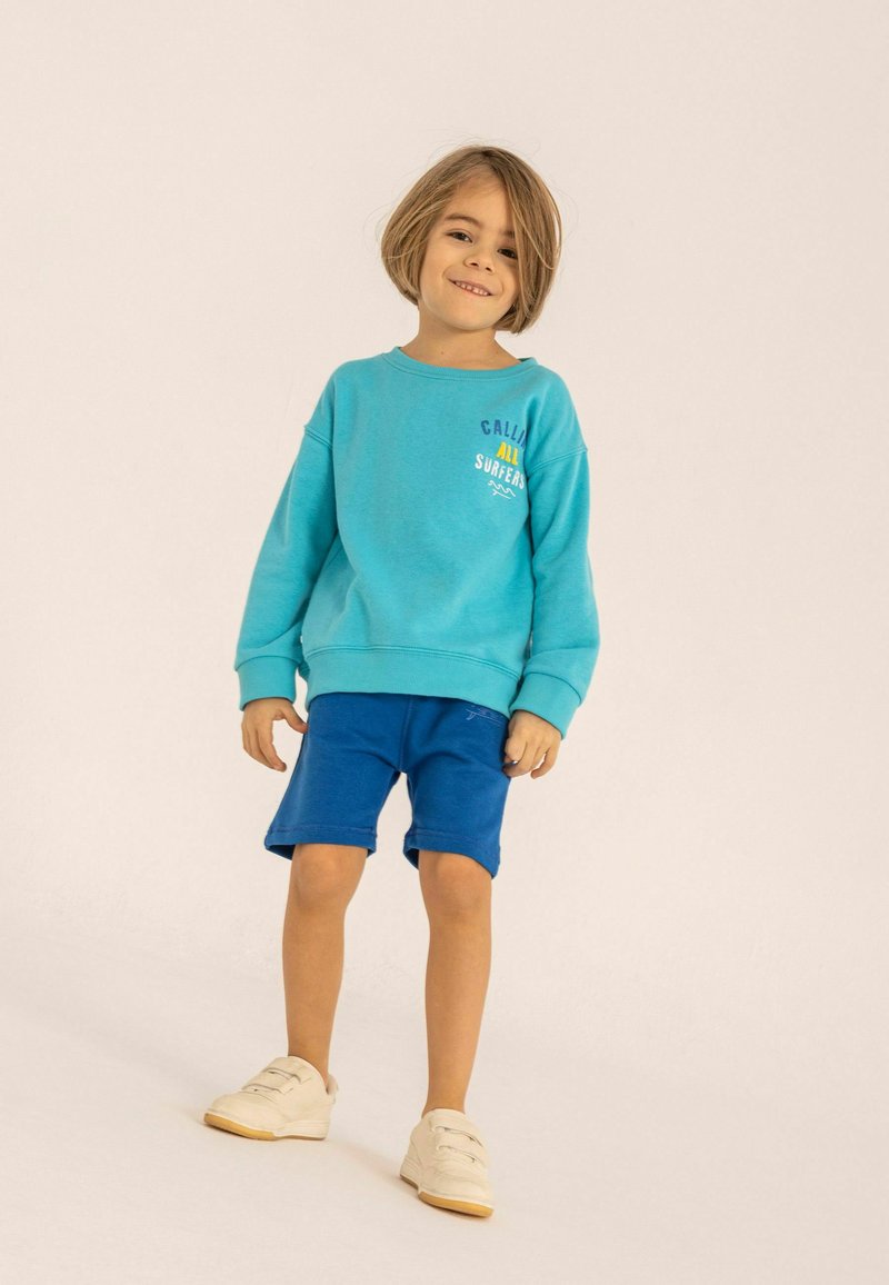 Ljus turkos sweatshirt med en logotyp, kombinerad med blå shorts. Barnet har på sig vita sneakers, vilket visar en avslappnad, casual outfit.