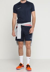 Man wearing navy blue Nike sportswear with white socks, a gray sweatshirt tied around waist, and bright orange and black running shoes.