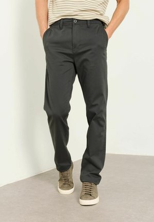 Mann trägt dunkelgraue Freizeithose mit Vordertaschen, kombiniert mit beigen Sneakers und einem gestreiften Hemd, steht auf einem hellen Boden.