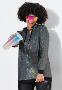 Hoodie met een zwarte basis bedekt met kleurrijke pailletten, die een glanzende, reflecterende textuur toont. Model houdt een gradient beker vast.