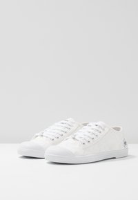 Baskets en toile blanches avec un motif texturé, semelle en caoutchouc blanc et lacets assortis. Elles possèdent un bout arrondi et un logo sur le côté.