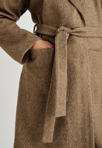 Abrigo de mezcla de lana marrón con cinturón atado, amplios bolsillos laterales y mangas anchas. La tela suave y texturizada ofrece una apariencia cálida.