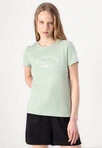 T-shirt en coton vert clair à manches courtes avec du texte blanc : "RIEN DE TEL QU'UN MAGNIFIQUE JOUR D'ÉTÉ." Associé à un short noir.