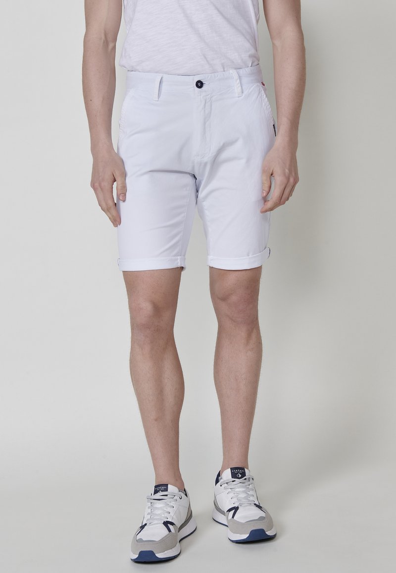 Homme portant un short blanc retroussé, un t-shirt blanc et des baskets blanches avec des détails gris et bleu marine, se tenant devant un fond uni.