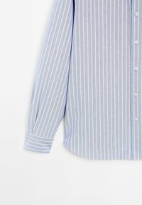 Jasnoniebieska koszula w poziome paski, wykonana z miękkiego materiału, z zapięciem na guziki z przodu, długimi rękawami i zaokrąglonym dołem. Posiada białe pionowe paski oraz standardowe mankiety.