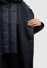 Črna podložena jakna z zadrgo spredaj, s karirasto notranjostjo, stranskimi žepi in kapuco, izdelana iz gladkega, vodoodpornega materiala.