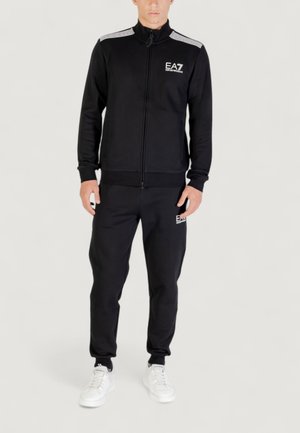 Tuta nera realizzata in tessuto morbido; giacca con zip e dettagli bianchi e logo sul petto, abbinata a pantaloni coordinati e sneakers bianche.