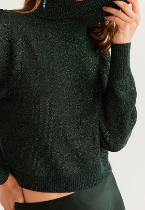 Kvinde iført en mørkegrøn glitrende rullekravebluse og sorte bukser, med synligt rødt hår og blå øreringe.