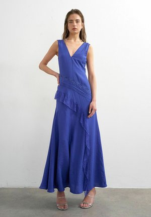 Femme en robe maxi bleue sans manches avec un détail de volants, debout, une main sur la hanche et portant des talons ouverts argentés.