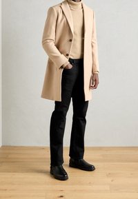 Abrigo largo color beige claro, suéter de cuello alto negro, jeans negros, botas de tobillo negras. Tejido de mezcla de lana, abrigo de un solo botón con dos botones.