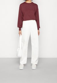 Osoba w bordowej bluzce z długim rękawem i białych, wysokich spodniach typu wide-leg, trzymająca białą torbę i nosząca białe sneakersy.