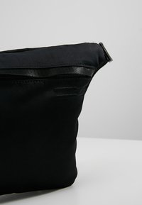 Sac banane en tissu noir avec fermeture éclair supérieure, forme incurvée et détail de sangle latérale réglable sur un fond gris uni.