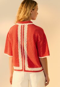 Röd öppenstickad topp med vit kant och geometriska mönster. Kortsleeves, rund halsringning och detalj på baksidan. Kombinerad med vita byxor.
