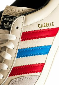 Fehér bőr sportcipő piros és kék csíkokkal, texturált díszítéssel, és fekete címkével, rajta az Adidas logóval. Arany "Gazelle" felirat az oldalon.