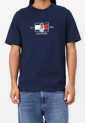 Marineblaue Baumwoll-T-Shirt mit kurzen Ärmeln, das ein grafisches Logo in Rot, Weiß und Schwarz zeigt. Der Text lautet "EST. 1985 HILFIGER."