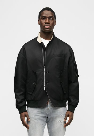 Homme portant une veste bomber noire zippée avec des poches à boutons et un jean gris délavé, debout devant un fond blanc uni.