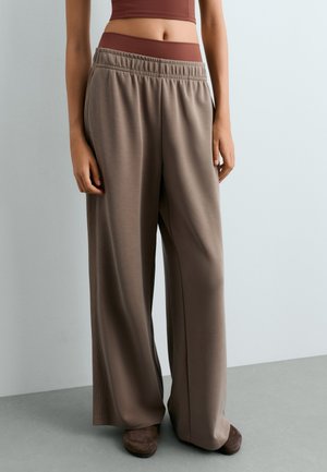 Personne portant un pantalon large marron ample, un crop top marron et des chaussures marron, debout sur un sol gris contre un mur gris clair.