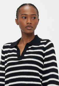 Kvinne med flette i håret iført en svart-hvitt stripete strikkegenser med svart krage, ser mot venstre mot en ensfarget bakgrunn.