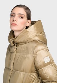 Puffig guldfärgad jacka med hög krage, blank yta och en logotyp på axeln. Har dubbel dragkedja och figursydd silhuett.