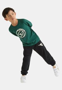 Maglietta di cotone verde con logo rotondo, pantaloni della tuta neri con accento bianco, abbinati a sneakers bianche. Ragazzo in posa dinamica.