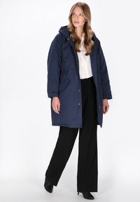 Marineblå lang parka med hætte, lynlås lukning, to sidelommer og afslappet pasform, båret over en lys skjorte og sorte bukser.