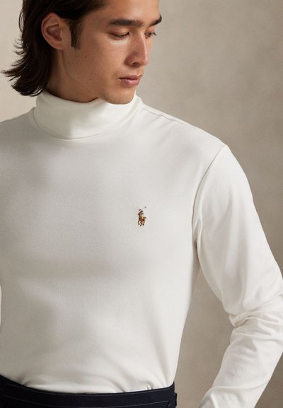 Jauns vīrietis baltā turtleneck džemperī ar mazu izšūtu polo spēlētāja logotipu uz krūšu daļas, skatās uz leju un uz sāniem.
