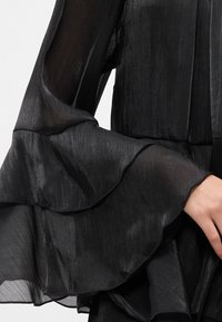 Must musta blouse pikad volangvarrukatega, tekstureeritud kangast ja sileda pinnaga, millel on õrn sära. Nähtav elegantne siluett.