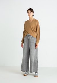 Tan innviklet genser i mykt strikkemateriale, kombinert med grå vid jeans og sølvfargede joggesko. Minimalistisk design, middels teksturer.