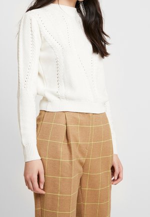 White knit sweater featuring a textured design with cable patterns and open-knit details. Paired with beige plaid trousers with yellow accents.