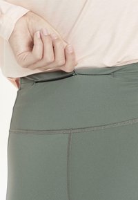 Korkeavyötäröiset vihreät leggingsit, joissa on sileä pinta, pienehkö vetskari-tasku ja vahvistetut saumat lisäämään kestoa.