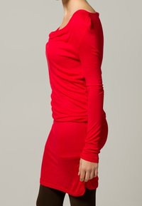 Rotes Langarmkleid mit Rundhalsausschnitt, lockerer Passform und gerafftem Stoff an der Schulter, kombiniert mit braunen Strumpfhosen. Weiches, dehnbares Material.