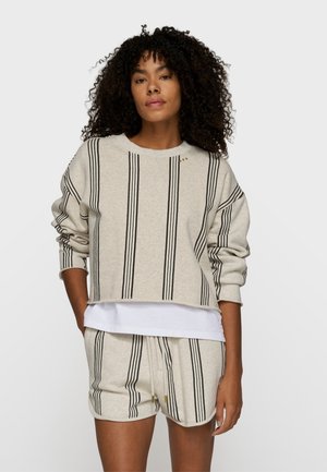 Frau mit lockigem Haar, die einen beigen Pullover mit langen Ärmeln und passende Shorts mit vertikalen schwarzen Streifen trägt, steht vor einem schlichten Hintergrund.