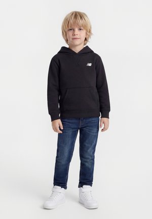 Bambino biondo in piedi, indossa una felpa con cappuccio nera New Balance, jeans blu e scarpe da ginnastica bianche su uno sfondo bianco uniforme.