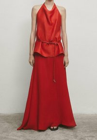Haut dos nu rouge avec une jupe longue fluide, présentant une coupe ample, un tissu texturé et un accent noué à la taille, créant un look superposé.