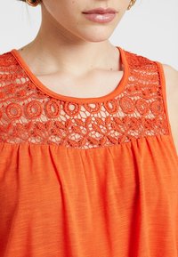 Pomarańczowy bezrękawnik z koronkową wstawką o okrągłych wzorach przy dekolcie, teksturowanym materiałem oraz marszczeniem na dole.