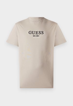 T-shirt di cotone beige a maniche corte, con un logo stampato "GUESS" e il testo "EST. 1981" in nero sulla parte anteriore. Design semplice.
