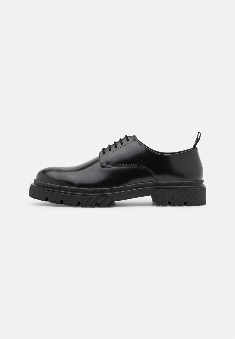 Chaussure derby en cuir noir poli avec semelle à crampons épaisse et cinq œillets, vue de côté sur fond blanc.