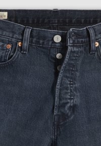 Jeans de mezclilla azul oscuro con cierre de botones, que presentan herrajes de metal y cinco bolsillos. La tela muestra una ligera textura y desvanecimiento.